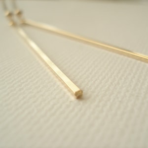 May include: A pair of long, thin gold earrings. The earrings are made of a square bar of metal.