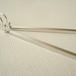 May include: A pair of silver-colored bar earrings. Each earring features a long, rectangular bar suspended from a small hook. The earrings are simple and minimalist in design, with a polished finish. The background is a light, textured surface.