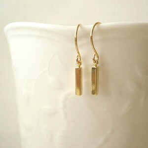 May include: A pair of gold-toned drop earrings featuring a small, vertical rectangular bar pendant suspended from a curved ear wire. The earrings have a polished, reflective metallic finish and a simple, clean design with no additional embellishments.