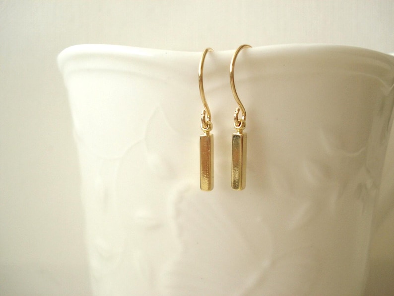 May include: A pair of gold-toned drop earrings featuring a small, vertical rectangular bar pendant suspended from a curved ear wire. The earrings have a polished, reflective metallic finish and a simple, clean design with no additional embellishments.