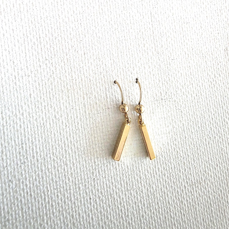 May include: A pair of gold-toned drop earrings featuring a small spherical bead at the top and a slender, rectangular bar pendant suspended from a curved ear wire. The earrings have a polished metallic finish and a simple, linear design.