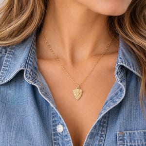 May include: A gold-toned necklace featuring a small, shield-shaped pendant with a textured surface and a cross design. The pendant hangs from a delicate, thin gold-toned chain that rests around the neck, providing a simple and refined appearance.