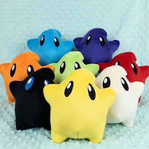 May include: A collection of eight plush star-shaped toys in various solid colours including yellow, black, orange, blue, purple, green, red, and white. Each soft star features two large, oval-shaped black eyes with white highlights, giving them a consistent appearance.