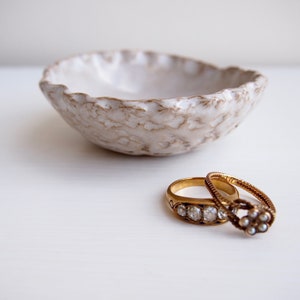 May include: Two gold rings with diamonds and pearls resting in a white ceramic bowl with a textured surface.