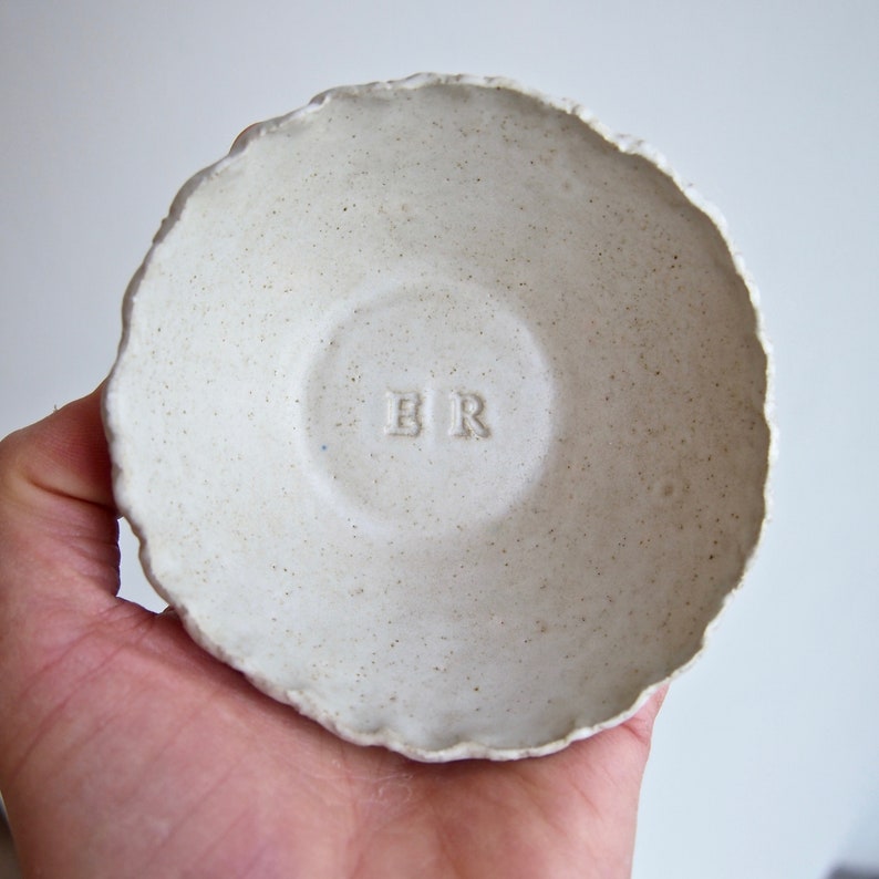 May include: A small, white ceramic bowl with a rough, textured surface. The bowl has a slightly scalloped edge and the initials "BR" are embossed on the bottom.