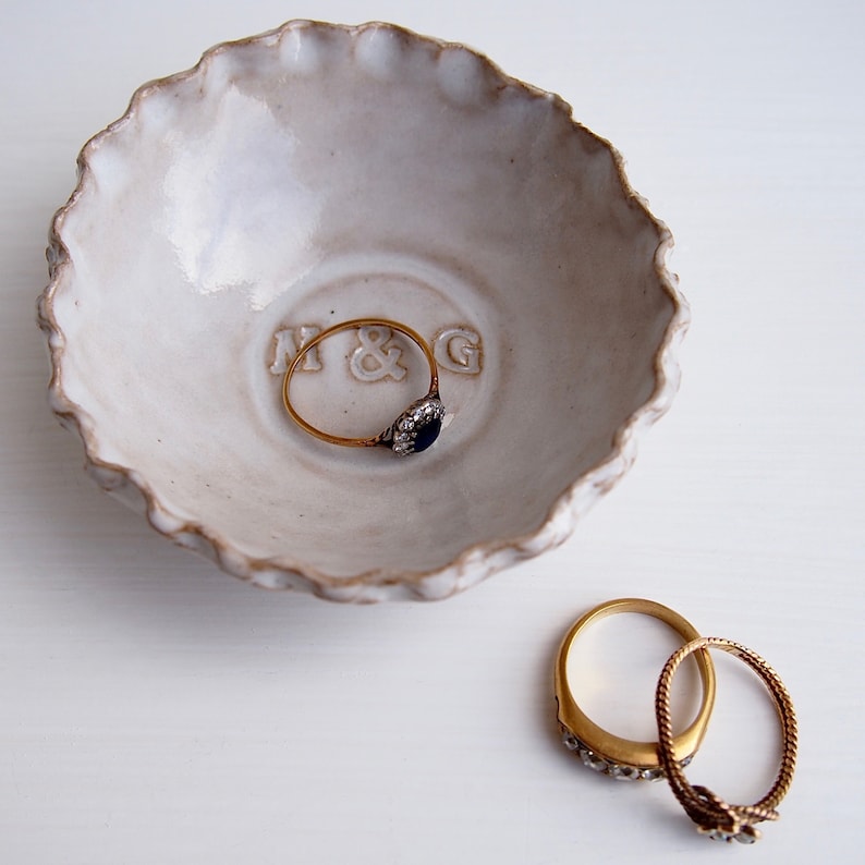 May include: White ceramic ring dish with a scalloped edge. The dish has the initials "M & G" embossed on the bottom. Two gold rings are sitting on a white surface next to the dish.
