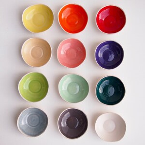 May include: Twelve small ceramic bowls in various colors, including yellow, red, pink, green, blue, purple, and white. The bowls are arranged in three rows of four, with the white bowl in the bottom right corner.