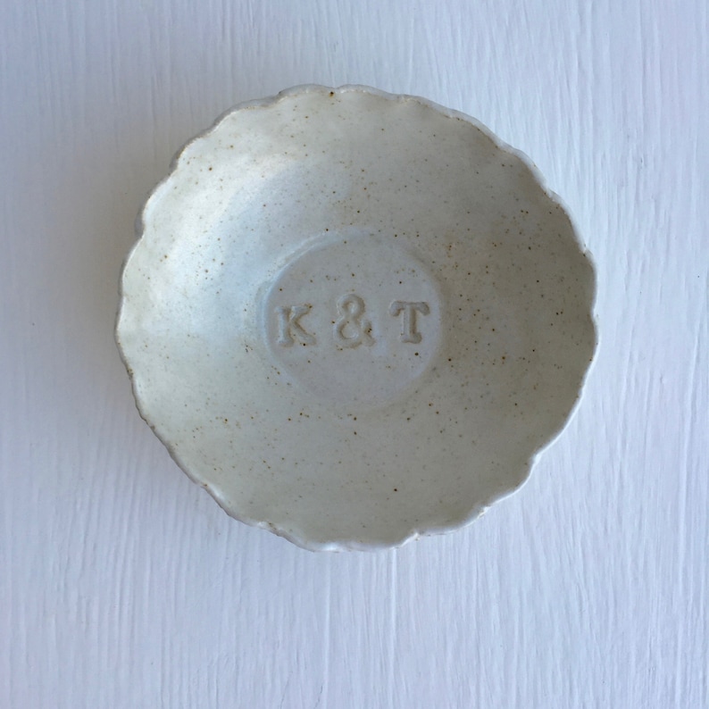 May include: A white ceramic ring dish with a scalloped edge. The dish has a circular imprint in the center with the letters "K & T" embossed in the center.