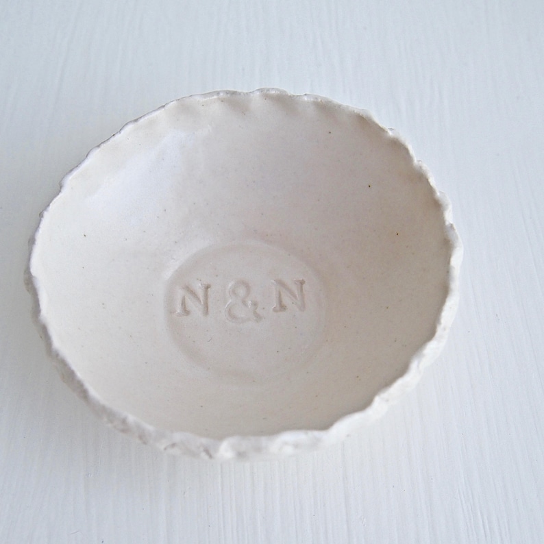 May include: White ceramic ring dish with a slightly rough texture and a raised inscription that reads "N&N".