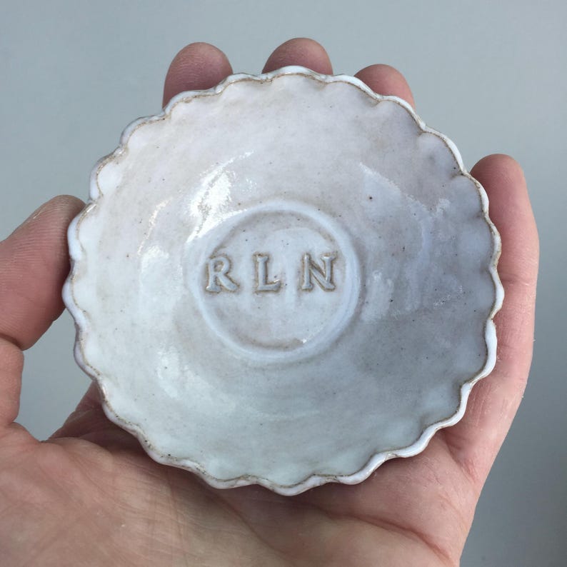 May include: White ceramic trinket dish with a scalloped edge. The dish has the initials "RLN" embossed in the center.