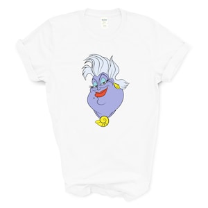 May include: White t-shirt with a cartoon image of Ursula from The Little Mermaid. Ursula is a purple sea witch with white hair and a mischievous expression.