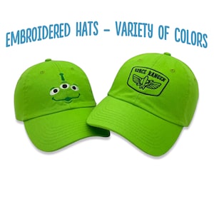 May include: Two green baseball caps with embroidered designs. One cap features a cartoon alien face with three eyes and a smile. The other cap has the text "Space Ranger" and a rocket ship with wings.