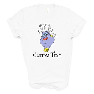 以下が含まれることがあります： 白いTシャツに、リトルマーメイドのウルスラの漫画の画像がプリントされています。画像の下には「CUSTOM TEXT」という文字がプリントされています。