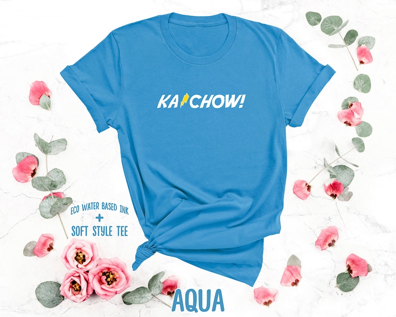 Op de afbeelding: Een lichtblauw T-shirt met de tekst "KACHOW!" in geel. Het shirt is gemaakt met milieuvriendelijke, watergebaseerde inkt en wordt beschreven als een zacht stijl T-shirt.