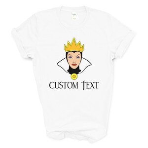 以下が含まれることがあります： 白いTシャツに、金色の冠と白黒の襟をつけた白雪姫の悪の女王の漫画の画像がプリントされています。画像の下には「CUSTOM TEXT」という文字が印刷されています。