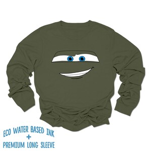 May include: A green long-sleeve shirt with a cartoon face printed on the front. The face has blue eyes and a white smile.