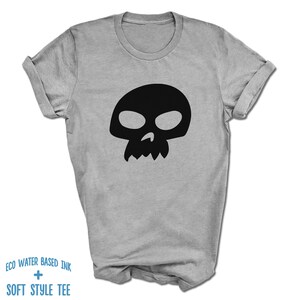 Pu&ograve; includere: Una maglietta grigia con una grafica di teschio nero. Il testo "ECO WATER BASED INK + SOFT STYLE TEE" &egrave; stampato sul fondo della maglietta.