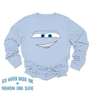 May include: Light blue long-sleeved t-shirt with a cartoon face of a car with blue eyes and a white outline. The text "ECO WATER BASED INK + PREMIUM LONG SLEEVE" is printed below the image.