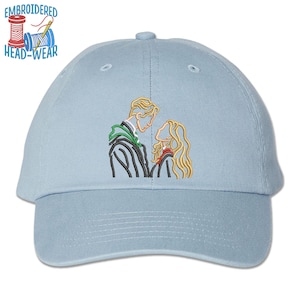 May include: Light blue baseball cap with an embroidered design of two figures in profile. The design features a man and woman with blonde hair, wearing green and red accents. The text "EMBROIDERED HEAD-WEAR" is above the cap.