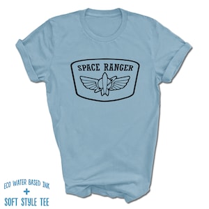 May include: Light blue t-shirt with a black graphic of a space ranger logo. The logo says "Space Ranger" and features a rocket ship with wings.