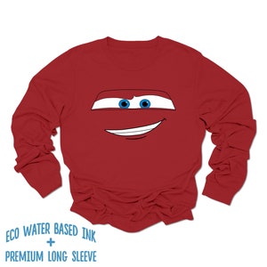 May include: Red long-sleeve shirt with a cartoon face of a red race car with blue eyes and a smile. The shirt has the text "ECO WATER BASED INK + PREMIUM LONG SLEEVE" printed on the bottom.