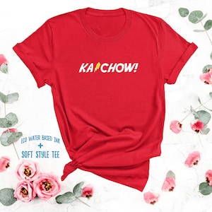 Op de afbeelding: Rood T-shirt met een witte bliksem en de tekst "KACHOW!" gedrukt op de voorkant. Het shirt is gemaakt met milieuvriendelijke, watergebaseerde inkt en een zachte stijl.