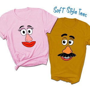 May include: Two t-shirts with a cartoon face design. The pink shirt has a female face with eyelashes and a red mouth. The gold shirt has a male face with a mustache and a red mouth. The text "Soft Style Tees" is in blue on a white background.