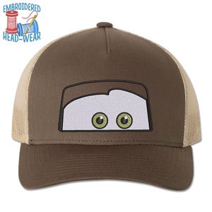 May include: Brown and tan trucker hat with an embroidered cartoon face with green eyes. The text "EMBROIDERED HEAD-WEAR" is above the face.