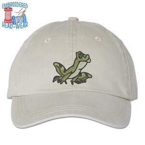 May include: A beige baseball cap with an embroidered green frog design. The cap has a curved brim and an adjustable strap.
