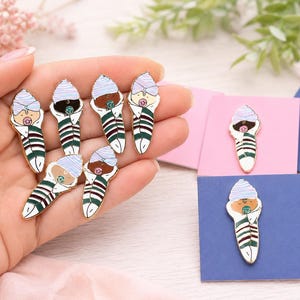 Pode incluir: Uma cole&ccedil;&atilde;o de pins de esmalte em forma de beb&eacute;s enrolados. Cada pin apresenta um beb&eacute; com um gorro &agrave;s riscas azul-claro e uma chupeta, enrolado numa manta &agrave;s riscas verdes, brancas e bordeaux. Os pins t&ecirc;m bordos de metal dourado.