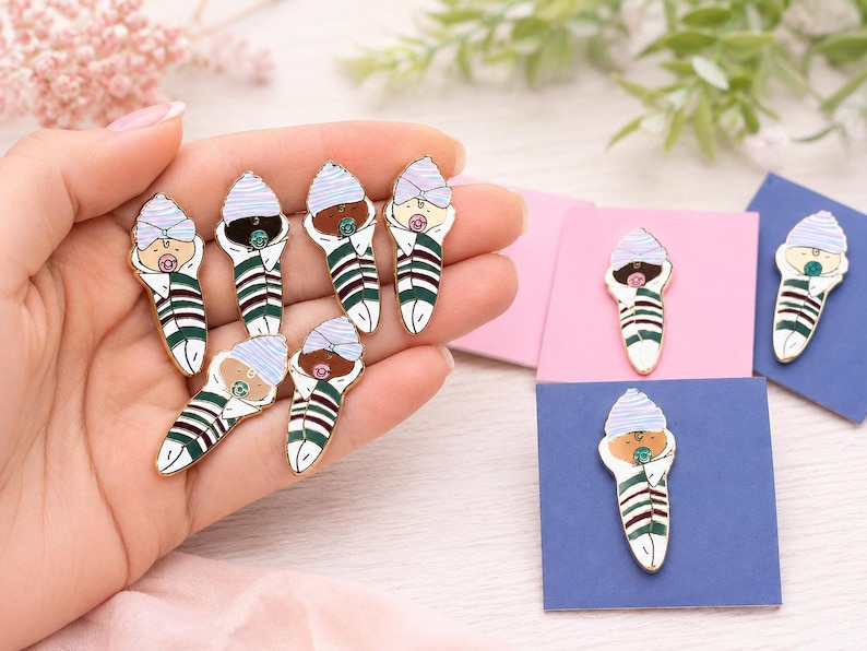 Pode incluir: Uma cole&ccedil;&atilde;o de pins de esmalte em forma de beb&eacute;s enrolados. Cada pin apresenta um beb&eacute; com um gorro &agrave;s riscas azul-claro e uma chupeta, enrolado numa manta &agrave;s riscas verdes, brancas e bordeaux. Os pins t&ecirc;m bordos de metal dourado.