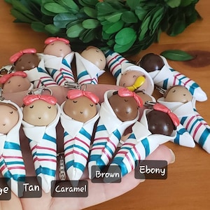 May include: A collection of 11 handmade polymer clay baby dolls, each wearing a white and blue striped swaddle. The dolls are arranged in a semi-circle on a hand, with the colors of the dolls' skin tones labeled below them: Beige, Tan, Caramel, Brown, Ebony.