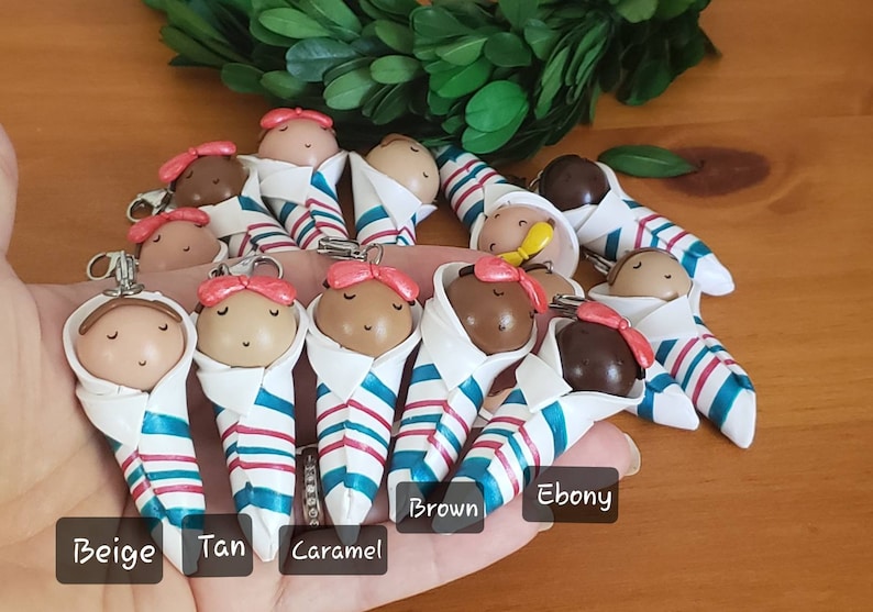 May include: A collection of 11 handmade polymer clay baby dolls, each wearing a white and blue striped swaddle. The dolls are arranged in a semi-circle on a hand, with the colors of the dolls' skin tones labeled below them: Beige, Tan, Caramel, Brown, Ebony.