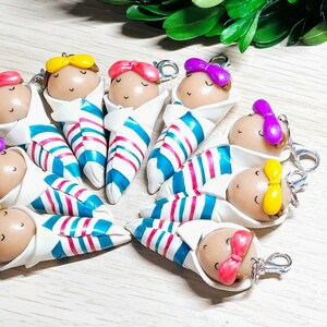 May include: A set of nine handmade polymer clay baby doll keychains. Each doll is wrapped in a blue and white striped blanket and has a different colored bow on its head. The keychains are perfect for adding a touch of whimsy to your keys, purse, or backpack.