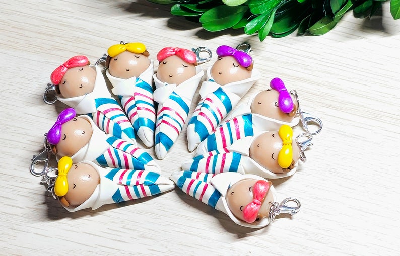 May include: A set of nine handmade polymer clay baby doll keychains. Each doll is wrapped in a blue and white striped blanket and has a different colored bow on its head. The keychains are perfect for adding a touch of whimsy to your keys, purse, or backpack.