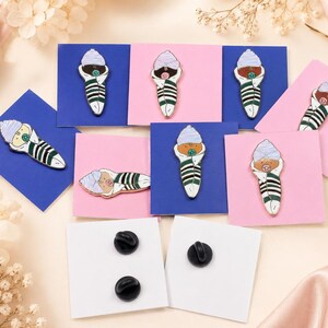 Pode incluir: Uma cole&ccedil;&atilde;o de pins de esmalte em forma de beb&eacute;s enrolados em mantas, com gorros e chupetas, exibidos em cart&otilde;es quadrados azuis e cor-de-rosa. Os pins apresentam mantas &agrave;s riscas verdes e brancas, com dois cart&otilde;es brancos a mostrar fechos pretos.