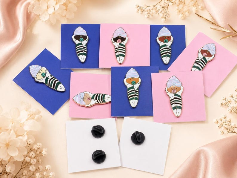 Pode incluir: Uma cole&ccedil;&atilde;o de pins de esmalte em forma de beb&eacute;s enrolados em mantas, com gorros e chupetas, exibidos em cart&otilde;es quadrados azuis e cor-de-rosa. Os pins apresentam mantas &agrave;s riscas verdes e brancas, com dois cart&otilde;es brancos a mostrar fechos pretos.