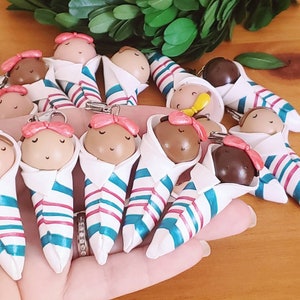 May include: A collection of 12 handmade polymer clay baby dolls, each wrapped in a white blanket with blue and pink stripes. The dolls have pink bows and closed eyes.