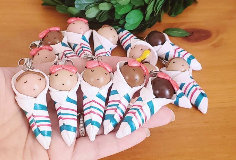 May include: A collection of 12 handmade polymer clay baby dolls, each wrapped in a white blanket with blue and pink stripes. The dolls have pink bows and closed eyes.