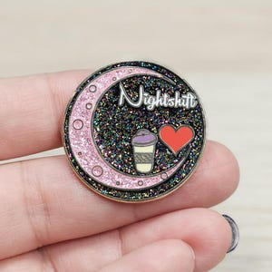 May include: A black and pink enamel pin with a crescent moon, a coffee cup, and a heart. The text "Nightshift" is written in white on the pin.