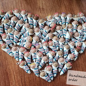 May include: A collection of handmade baby doll ornaments, each wrapped in a blue and red striped blanket. The dolls have different colored hair and skin tones. The text "Handmade to order" is visible in the bottom right corner of the image.