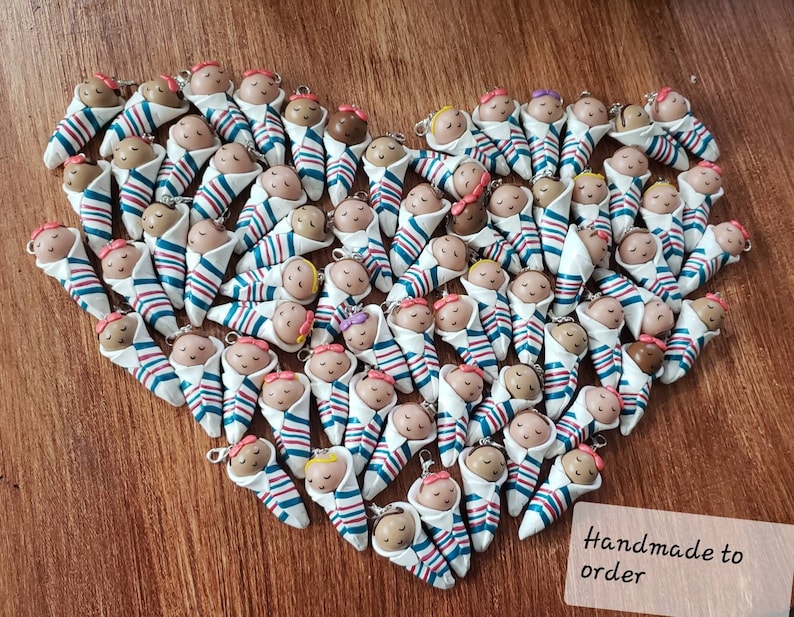 May include: A collection of handmade baby doll ornaments, each wrapped in a blue and red striped blanket. The dolls have different colored hair and skin tones. The text "Handmade to order" is visible in the bottom right corner of the image.