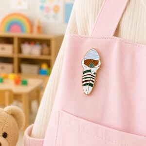 Pode incluir: Um broche de esmalte met&aacute;lico em forma de beb&eacute; enrolado numa manta, usando um gorro &agrave;s riscas azul-claro e branco. O beb&eacute; tem pele castanha, uma chupeta verde e est&aacute; enrolado numa manta branca com riscas horizontais verde-escuras, vermelhas e brancas.