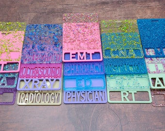 May include: A set of ten colorful glitter badge holders with different medical professions printed on them. The professions include PT, OTA, OT, Ultrasound, X-ray, Radiology, EMT, Pharmacy, ED Physician, CNA Instructor, RT, LPN, ARNP, and RN.