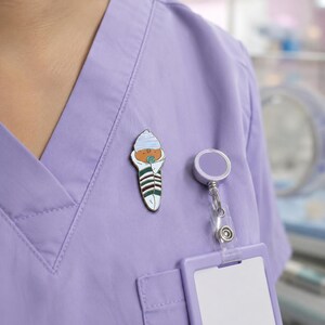 Pode incluir: Um broche de esmalte met&aacute;lico em forma de beb&eacute; enrolado num cobertor, usando um gorro &agrave;s riscas azuis e brancas. O beb&eacute; est&aacute; embrulhado numa manta branca com riscas horizontais verde-escuras e bordeaux e tem uma pequena chupeta azul.