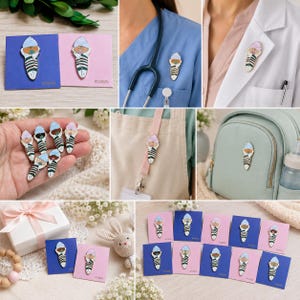 Pode incluir: Um alfinete de esmalte met&aacute;lico com um beb&eacute; enrolado numa manta e uma chupeta, dispon&iacute;vel com gorro azul claro ou rosa claro. O beb&eacute; est&aacute; envolvido numa manta branca com riscas verde-escuras em pequenos cart&otilde;es quadrados.