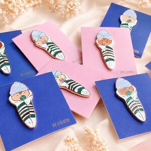 Pode incluir: V&aacute;rios pins de esmalte em forma de beb&eacute; enrolado numa manta, com um gorro &agrave;s riscas azuis e brancas, uma manta &agrave;s riscas verdes e brancas e uma chupeta. Cada pin est&aacute; preso a um cart&atilde;o retangular com a inscri&ccedil;&atilde;o VV's Grotto em cartolina azul ou rosa.