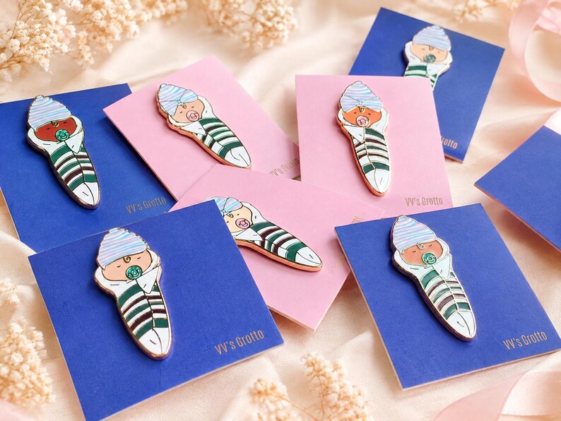 Pode incluir: V&aacute;rios pins de esmalte em forma de beb&eacute; enrolado numa manta, com um gorro &agrave;s riscas azuis e brancas, uma manta &agrave;s riscas verdes e brancas e uma chupeta. Cada pin est&aacute; preso a um cart&atilde;o retangular com a inscri&ccedil;&atilde;o VV's Grotto em cartolina azul ou rosa.
