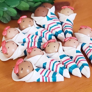May include: Twelve small, handmade clay figurines of babies wrapped in white blankets with teal and pink stripes. The babies have brown skin and pink bows on their heads.
