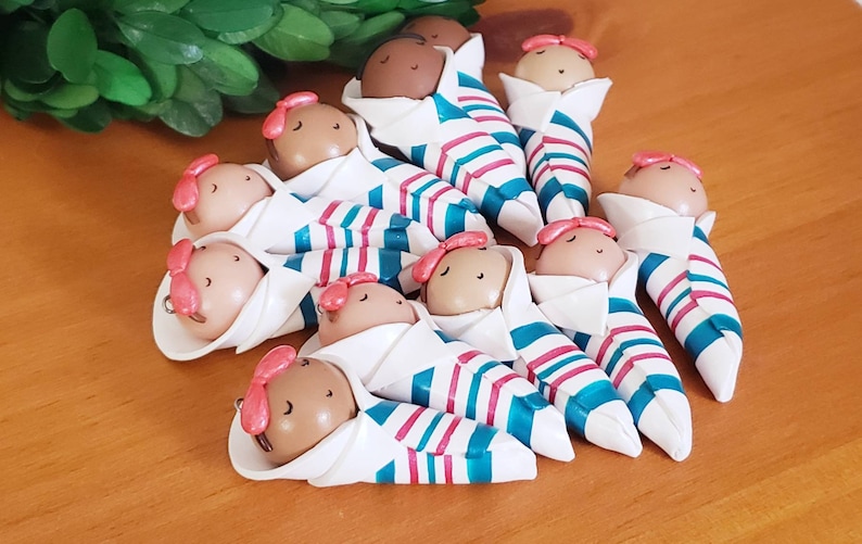 May include: Twelve small, handmade clay figurines of babies wrapped in white blankets with teal and pink stripes. The babies have brown skin and pink bows on their heads.